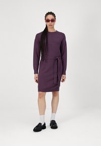 Violet megztinis su ilgomis rankovėmis, liemens juostele ir apvaliu iškirpimu. Modelis dėvi juodus batus ir baltas kojines.