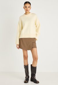 Κρεμ πουλόβερ με ριμπ λαιμόκοψη, συνδυασμένο με καφέ φούστα σε σχέδιο houndstooth. Το μοντέλο φοράει καφέ δερμάτινες μπότες μέχρι το γόνατο με διακοσμητικές αγκράφες.