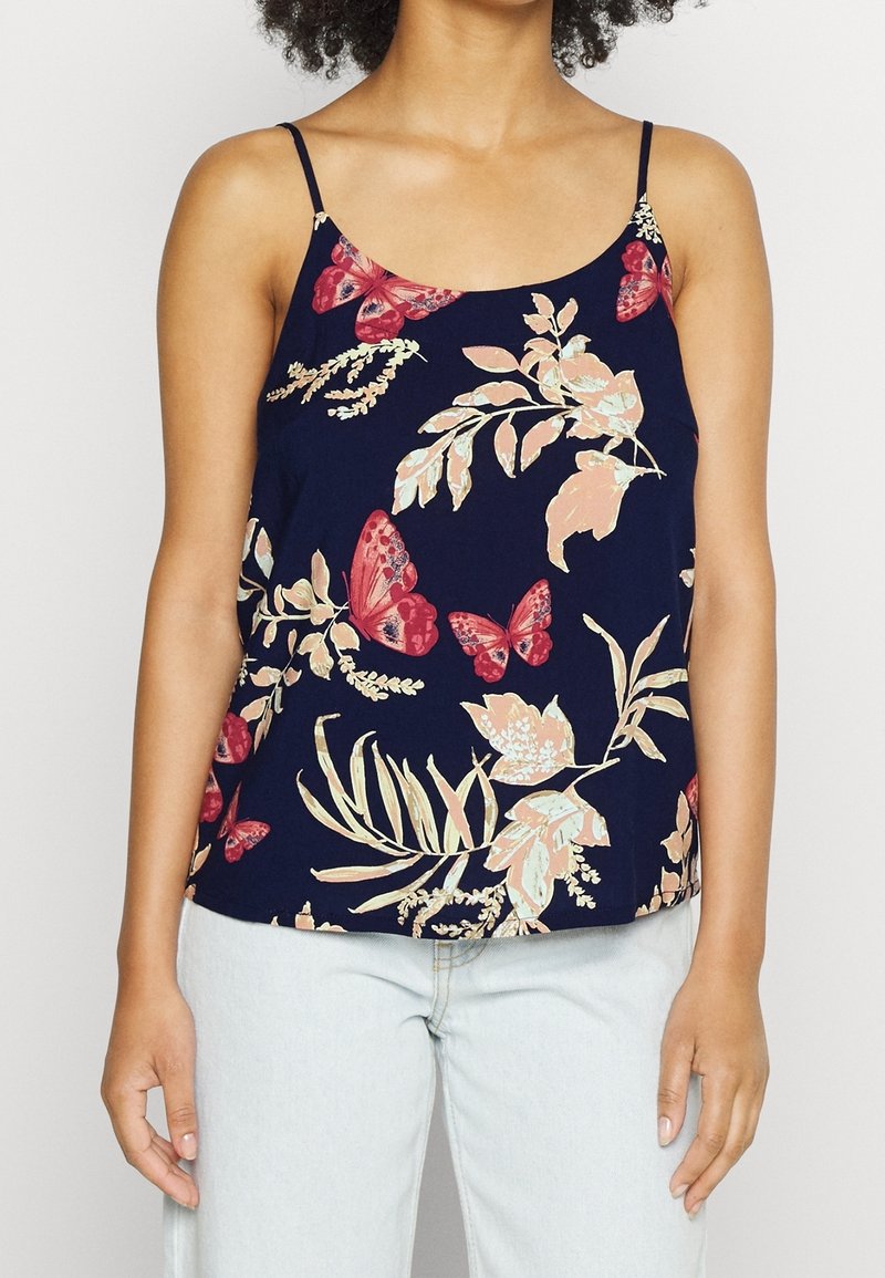 Femme portant un caraco bleu marine avec des papillons rouges et un imprimé floral beige, associé à un jean bleu clair.