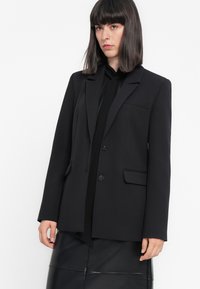 Donna con capelli lunghi e lisci di colore scuro, che indossa un blazer nero sopra una blusa nera e una gonna di pelle nera, su uno sfondo bianco uniforme.