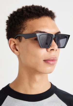 Crne kutne sunčane naočale s tamnim lećama, koje imaju hrabar geometrijski dizajn i mat finiš. Nose ih osoba s kratkom kosom.