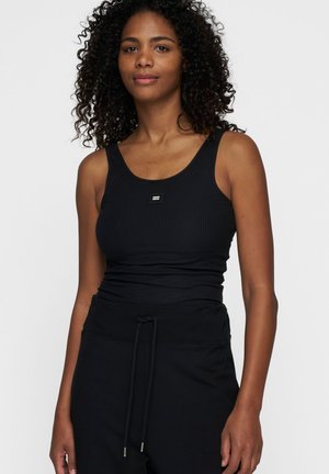 Donna con capelli ricci, che indossa un top a coste nero e pantaloni neri con coulisse, in piedi su uno sfondo bianco uniforme.