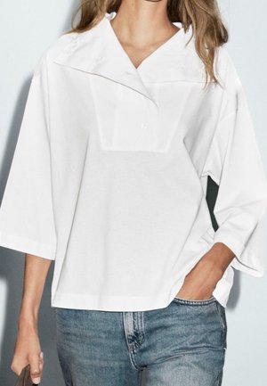 Femme portant un chemisier blanc ample à manches larges et col large, associé à un jean bleu. Une main dans la poche, l'autre tenant un sac.
