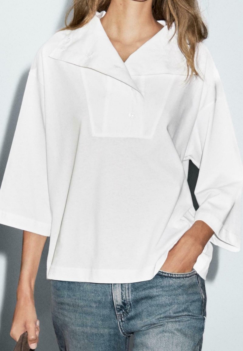 Femme portant un chemisier blanc ample à manches larges et col large, associé à un jean bleu. Une main dans la poche, l'autre tenant un sac.