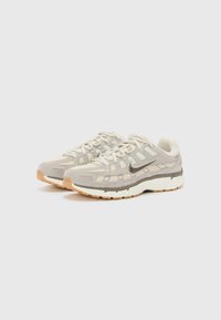 Beżowe sneakersy sportowe z szarymi siateczkowymi wstawkami, okrągłym noskiem, białą podeszwą z teksturowanym gripem oraz wyraźnym systemem sznurowania. Logo marki z boku.