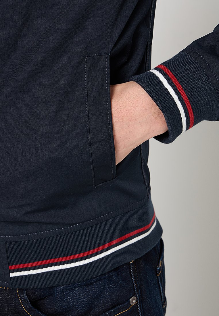 Veste bleu marine avec un ourlet côtelé orné de rayures rouges, blanches et bleues ; comprend une poche latérale et un tissu lisse et texturé.
