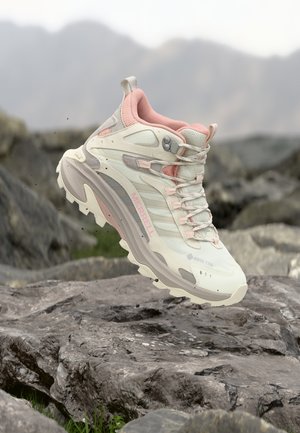 Lichtbeige en roze Merrell wandelschoen met robuuste zool zwevend boven rotsachtig terrein, berglandschap wazig op de achtergrond.