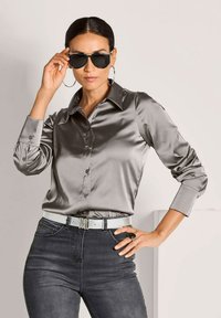 Frau in glänzender grauer Satinbluse und dunklen Jeans, die schwarze Sonnenbrille anpasst und große Hoop-Ohrringe, einen silbernen Gürtel und einen schwarzen Ring trägt.