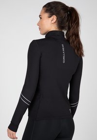 Schwarzes langärmeliges Sportoberteil mit hohem Kragen, das reflektierende Streifen an den Ärmeln und ein aufgedrucktes Logo auf dem Rücken hat.