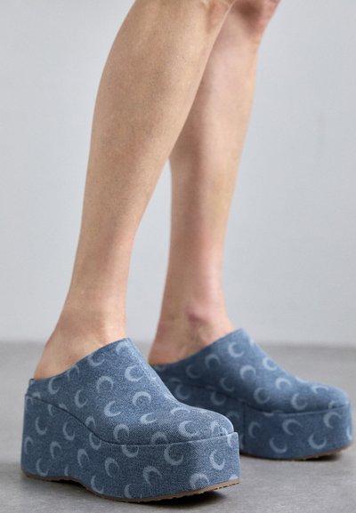 Sabots plats bleus avec une texture douce semblable à du denim, présentant un motif répétitif de croissants de lune bleu clair et une semelle en caoutchouc épaisse.