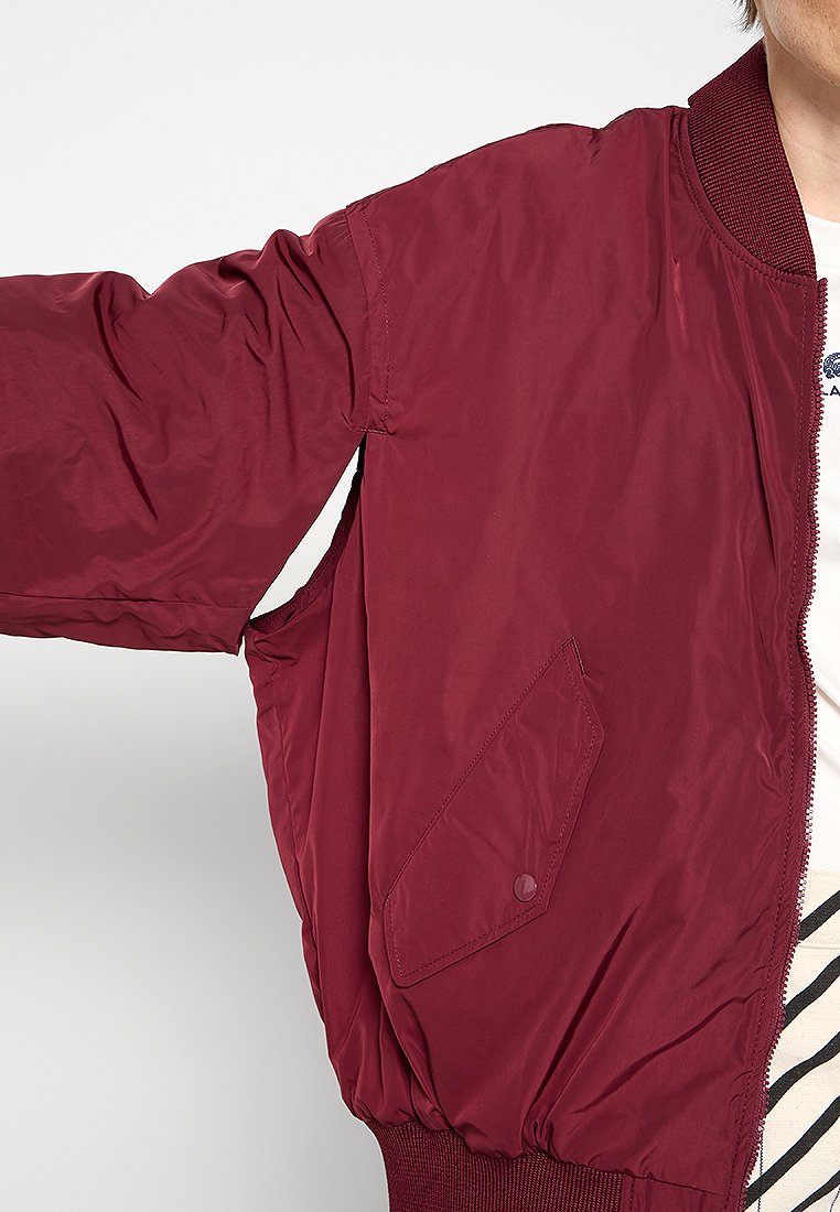 Veste bomber bordeaux en nylon lisse, avec des poignets côtelés et une poche avant avec bouton pression.