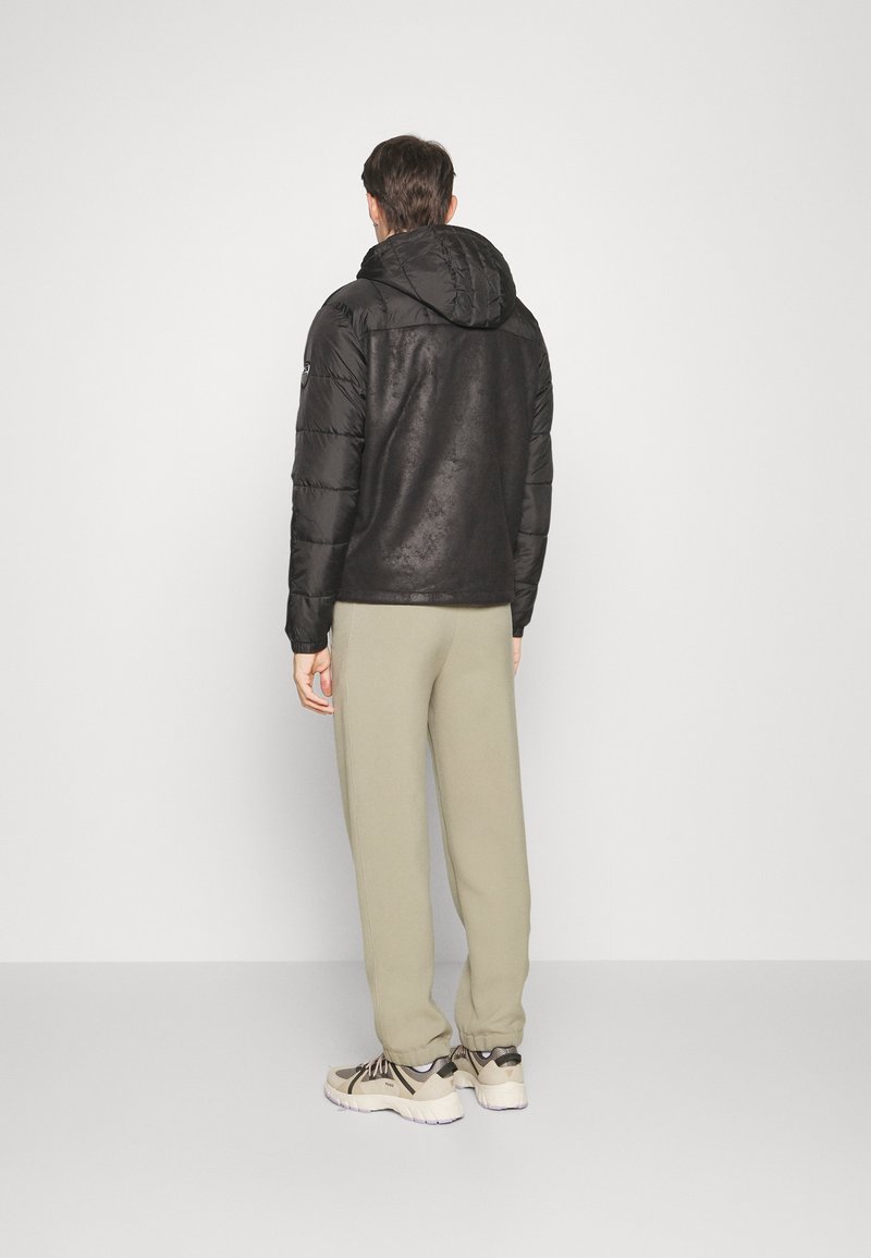 Svart pufferjacka med huva och matt yta, i kombination med ljusbeige joggers och beiga sneakers, sedd bakifrån.