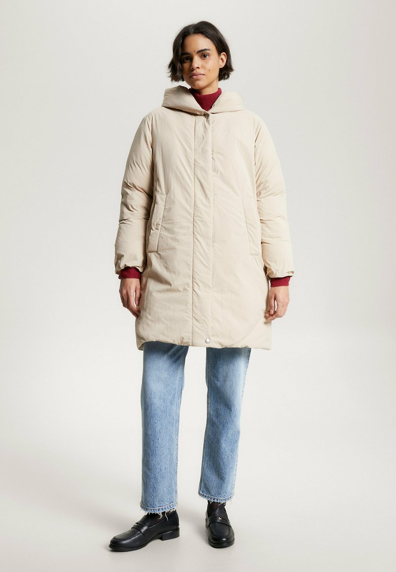 Beige Daunenjacke mit Kapuze, vorderer Knopfleiste und Seitentaschen; kombiniert mit hellblauen Jeans und schwarzen Mokassins. Glatte Textur.