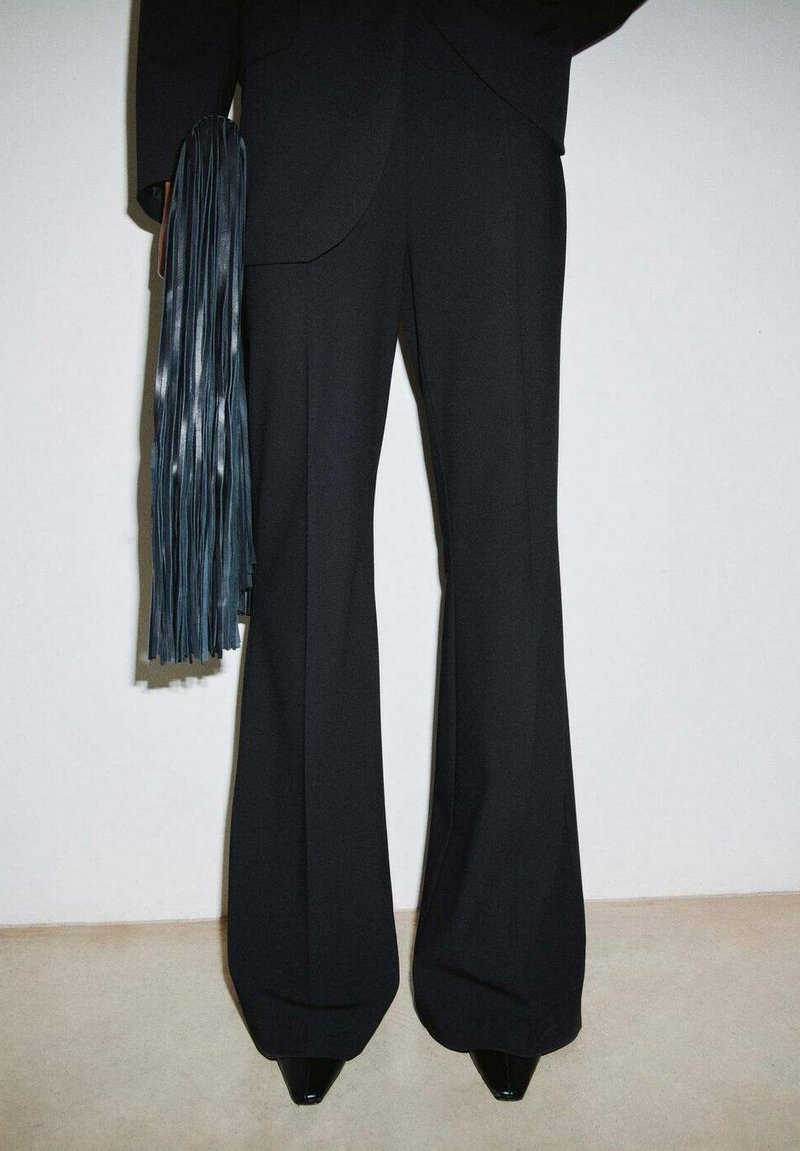 Pantaloni neri su misura con silhouette svasata, dotati di una texture liscia, vita alta e abbinati a un accessorio turchese con frange.