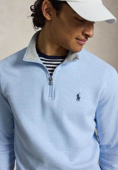 Mlada oseba nosi svetlo modro pulover z zadrgo in majhno temno modro logo igralca polot, pod njim črtasto srajco in belo kapo, gleda navzdol.