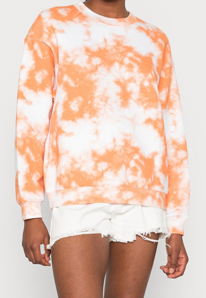 Personne portant un sweat-shirt tie-dye orange et blanc et un short en denim blanc effiloché, debout devant un fond uni.