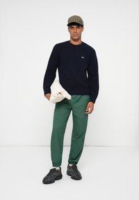 Tumesinine kudum sweaters, rohelised elastse vöökohaga püksid, hallid tossud ja ruuduline müts. Modell hoiab valget rõivast, seistes ühtlase tausta ees.