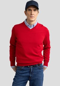 Crvena v-neck džemper od pletenog materijala, uparena s plavom i bijelom prugastom košuljom s kragnom i plavim trapericama. Jednostavan, prilagođen dizajn.