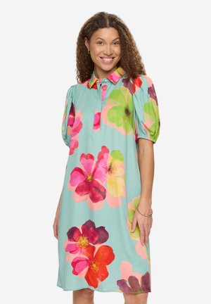 Kvinde smilende i en knælængde, lyseblå kjole med store farverige blomsterprint og korte pufærmer, stående foran en ensfarvet baggrund.