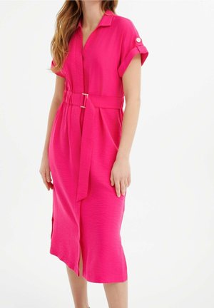 Mujer con un vestido midi rosa brillante, de manga corta, ajustado con cinturón y con cuello, de pie sobre un fondo blanco liso.