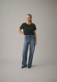 Naine seisab käsi puusal, seljas must t-särk, kõrge vöökohaga laiade sääreosadega sinised teksad ja mustad tippudega kingad, taust on lihtne.