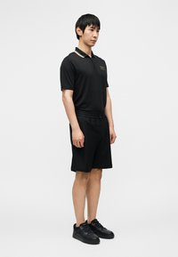 Μαύρο πόλο πουκάμισο με γιακά διακοσμημένο με χρυσές λεπτομέρειες, συνδυασμένο με μαύρο σορτς και μαύρα αθλητικά παπούτσια. Απλός, καθαρός σχεδιασμός με εφαρμοστή γραμμή.