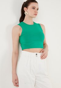Zelený bezrukávový crop top vyrobený z elastického materiálu, s okrúhlym výstrihom, kombinovaný s bielymi džínsovými nohavicami s vysokým pásom.