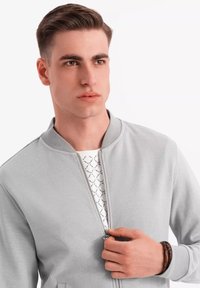 Cazadora bomber de color gris claro con cuello de canalé y cremallera en el frente. Debajo, se asoma una camisa blanca con un patrón; muñeca adornada con una pulsera marrón.