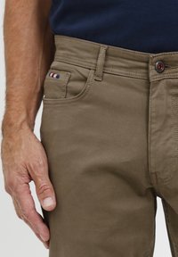 Spodnie khaki o gładkiej teksturze, z przednią kieszenią ozdobioną niebiesko-biało-czerwonymi paskami i czerwonym guzikiem w pasie.