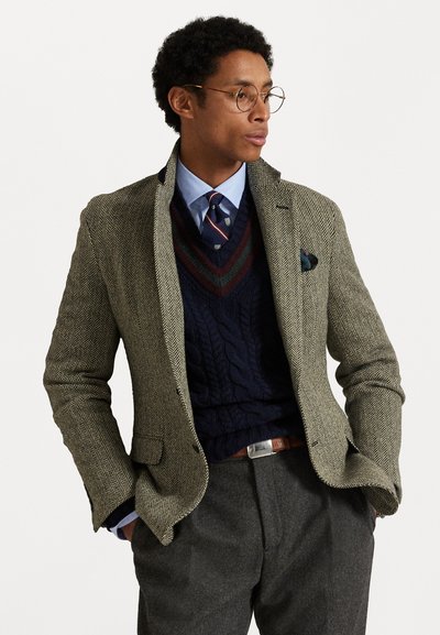 Blazer em padrão espinha de peixe em verde oliva, sobre um suéter de malha azul-marinho, com uma camisa azul clara e uma gravata estampada, complementado por calças cinzas.