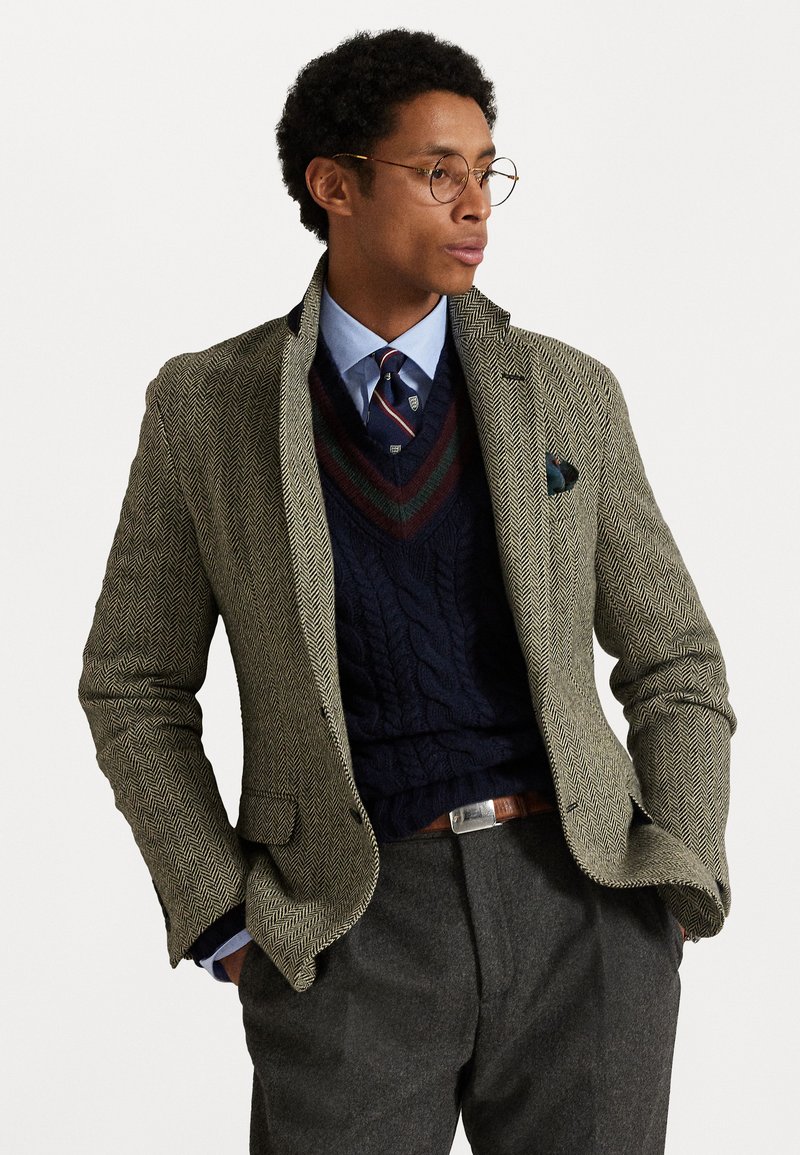 Blazer a zig zag in verde oliva, indossato sopra un maglione a trecce color navy, con una camicia azzurra e una cravatta stampata, completato da pantaloni grigi.