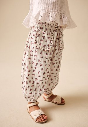 Kind draagt witte losse broek met bloemenpatroon en elastische manchetten, een wit gestructureerd topje met ruches aan de zoom en crème sandalen met enkelbandjes.