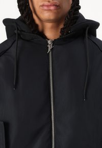 Black zip-up jacket with a matte finish, featuring a hood, silver hardware, and elastic accents on cuffs and neckline. Textured fabric visible.