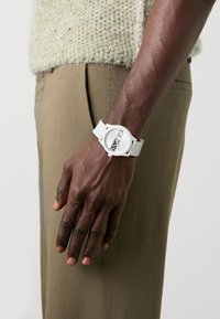 Montre en caoutchouc blanche avec un cadran rond arborant le logo "GUESS" en noir. Portée sur une main à la peau foncée, accompagnée d'un pantalon de couleur claire.