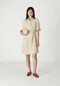 Mulher vestindo um vestido bege com cinto, segurando uma clutch a condizer e sapatos rasos vermelhos, em pé contra um fundo claro e liso.