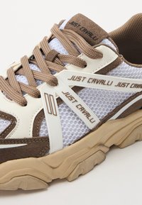Sneaker med en hvit mesh-overdel og brune semskede detaljer, teksturerte snøringer og en kraftig beige såle; inkluderer merkevaredetaljer i brunt.