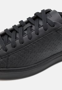 Baskets noires en cuir texturé, avec des motifs géométriques en relief, des lacets plats noirs et une semelle en caoutchouc lisse.