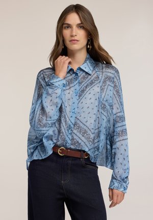 Frau mit langen braunen Haaren, die ein halbtransparentes blau gemustertes Hemd mit Knöpfen, dunkle Jeans und einen braunen Gürtel vor neutralem Hintergrund trägt.