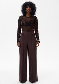 Vrouw staat gekleed in een doorschijnend zwart bloemenbloesje met lange mouwen over een zwarte beha, hooggeknoopte brede bruine broek, gouden hoopsieraden en armbanden.