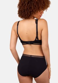 Mujer con cabello rizado que lleva un sujetador negro de encaje y ropa interior de cintura alta a juego, vista desde atrás sobre un fondo liso.