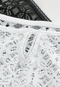 Tissu en dentelle florale noir et blanc avec des motifs complexes et des bords festonnés se superposant sur un fond blanc.