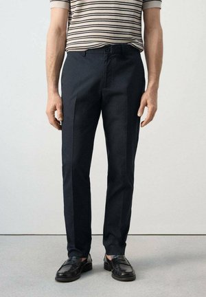 Mann trägt schwarze, maßgeschneiderte Hosen, schwarze Lederloafers und ein beige-schwarz gestreiftes Kurzarmhemd, steht vor einem schlichten Hintergrund.