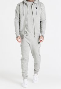 Szary zestaw bluzy z kapturem i dresów wykonany z miękkiego materiału, z przodem na zamek błyskawiczny, bocznymi kieszeniami i haftowanymi detalami logo, w połączeniu z białymi trampkami.