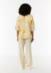 Kurzärmlige Bluse in hellgelb mit abstrakten Mustern in Beige und Gelb; kombiniert mit cremefarbenen, weit geschnittenen Hosen.