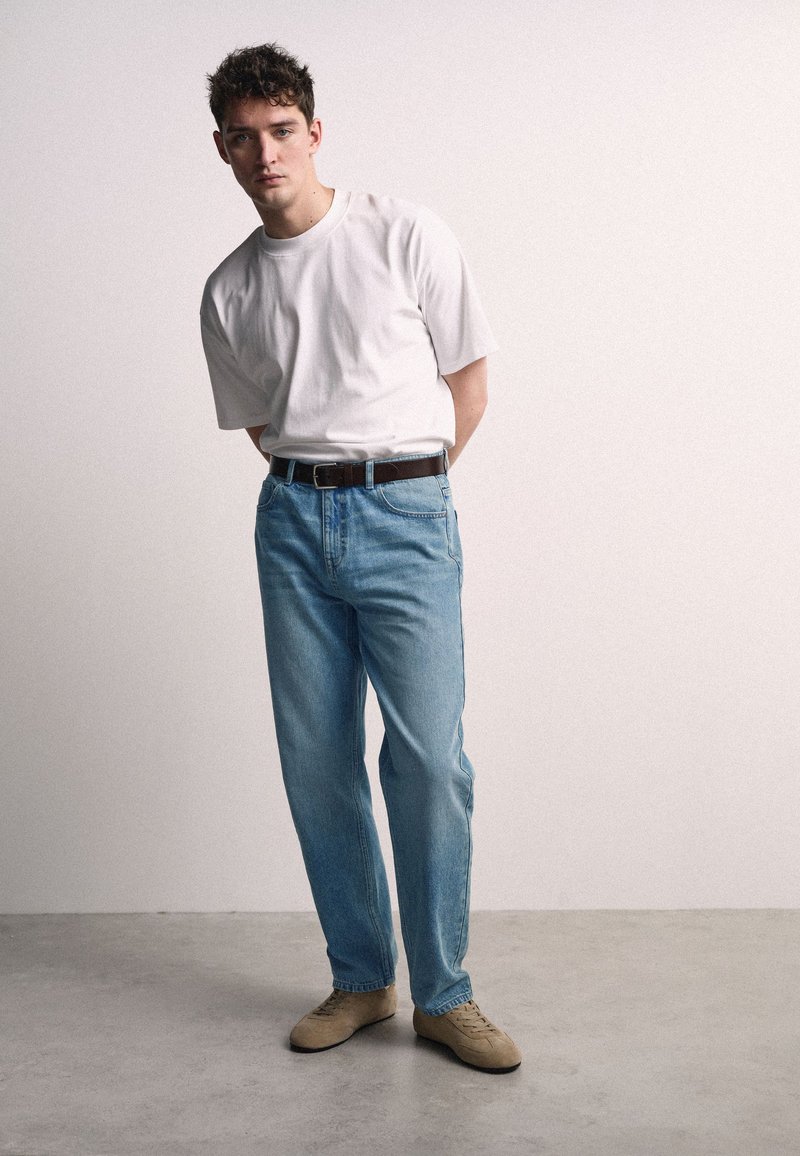 Jonge man staat met handen achter zijn rug, draagt een wit T-shirt, lichtblauwe spijkerbroek, bruine riem en beige sneakers op een grijze vloer.