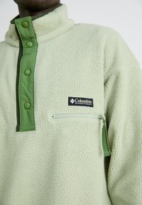 Fleece-pullover i lysegrønn med glidelås-lomme på brystet, grønn trykknappslisse og Columbia-logo-patch på brystet. Myk tekstur.