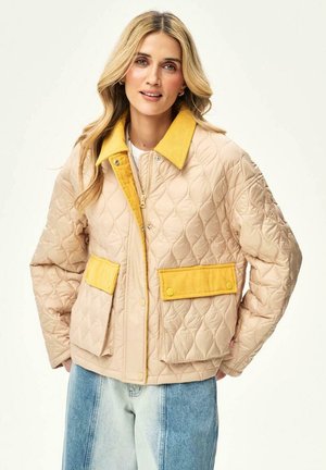 Blondynka ubrana w beżową pikowaną kurtkę z żółtym kołnierzem i patkami na kieszeniach, w połączeniu z dwukolorowymi niebieskimi dżinsami, na jednolitym tle.