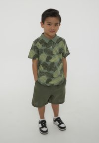 Polo shirt a maniche corte in khaki con motivo a foglie verdi, abbinato a pantaloni corti oliva. Calzature: sneakers nere e bianche.