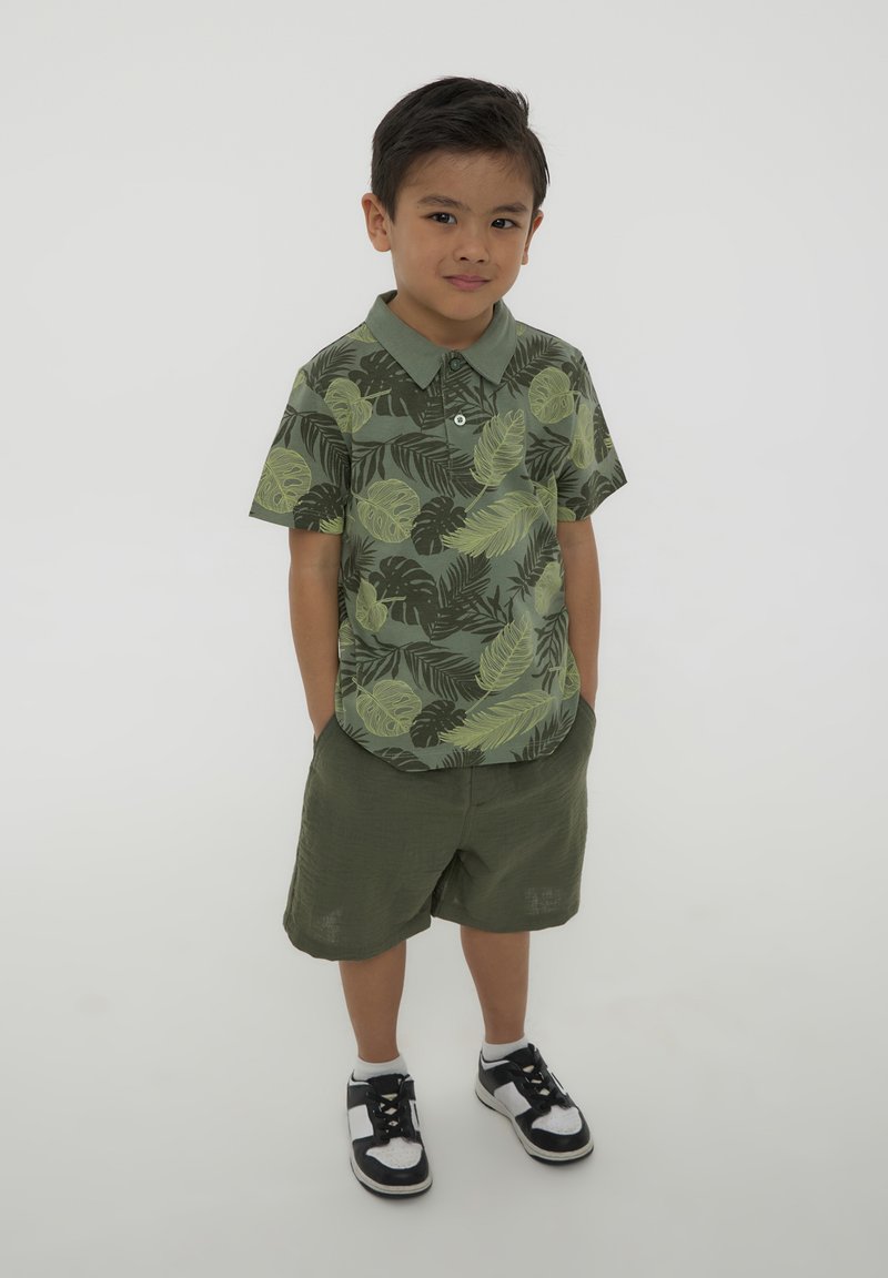 Polo shirt a maniche corte in khaki con motivo a foglie verdi, abbinato a pantaloni corti oliva. Calzature: sneakers nere e bianche.