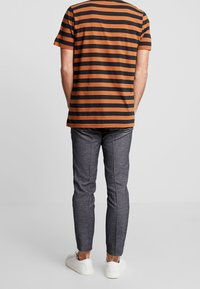 Hombre de pie, de espaldas, vistiendo una camiseta de manga corta a rayas naranja y negra, pantalones grises y zapatillas blancas sobre un fondo liso.